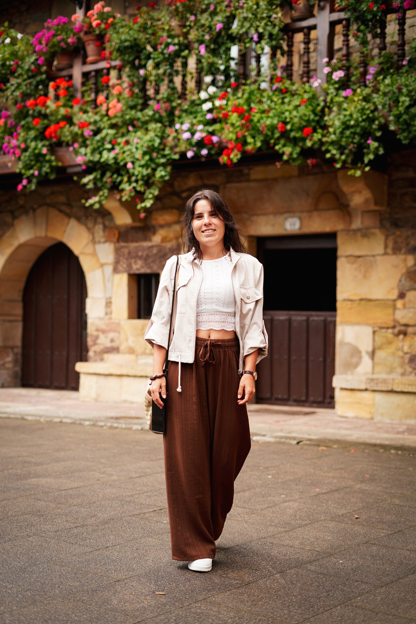 A stylish young woman wearing a white lace crop top, light beige jacket, and brown wide-leg trousers
