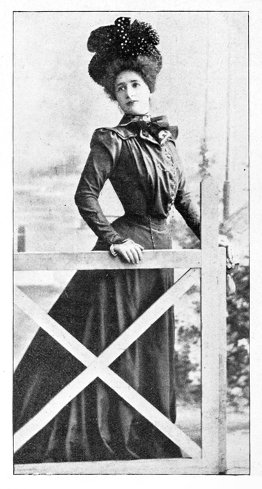 A black and white photographic print depicting a woman in a large, ornate hat and period dress, leaning casually on a wooden fence outdoors. This 19th-century image captures the fashion and posture typical of the era's portraiture
