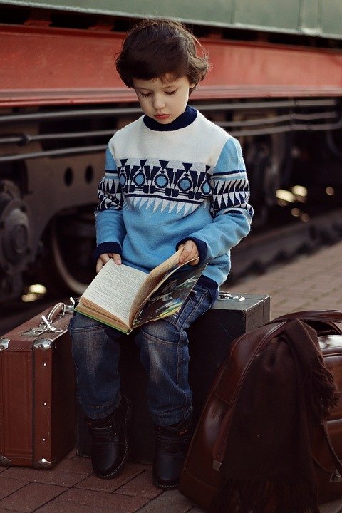 A boy reading a book by the side of the train while sitting on suitcases