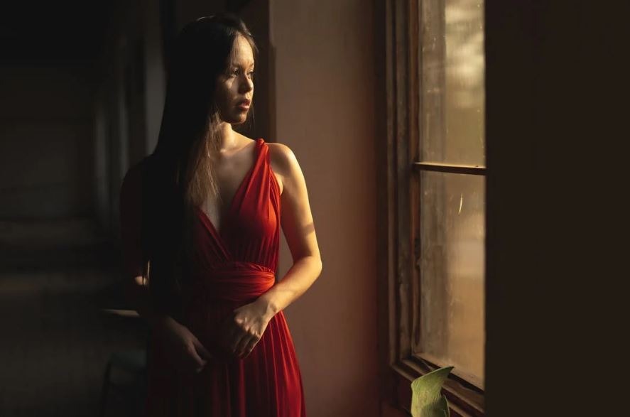 a woman in a dark wearing a red bias cut dress looking towards the window