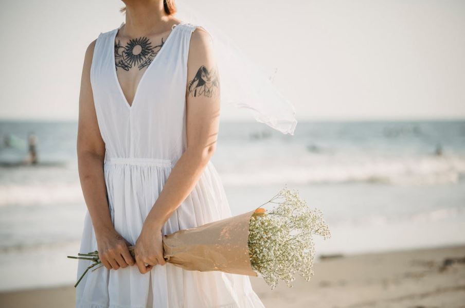a woman wearing a white bias cut dress and holding flowers at the beach